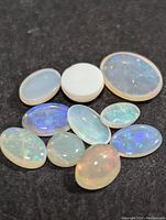 Ten genuine opal gemstones displayed on black surface, showing variety of oval shapes and play-of-color.