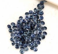 Close-up photo showing multiple small round faceted sapphires in different blue hues spread out on a white surface.