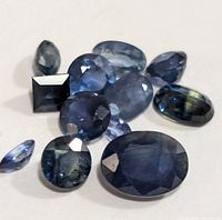 Close-up of multiple genuine sapphire gemstones showing oval, round, and rectangular faceted cuts in varied blue tones.