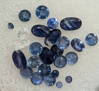 Top-down photo showing various loose blue sapphires and one clear gemstone on a white background with different cuts and shades of blue.