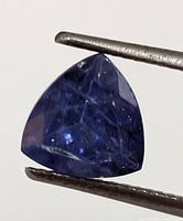 Front view of triangular cut tanzanite held by tweezers showing rich blue-violet color and faceting detail.