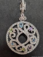Front and angled views of a round silver pendant with intricate openwork filigree design with small clear stones, blue topaz, amethyst, and peridot accent stones.