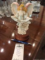 Front view of the musical angel figurine showing the 'Gloria' banner and the wooden base, pen for scale.