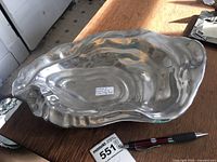 Photo of the oyster-shaped aluminum tray on a wooden surface with pen and maxsold tag for scale.