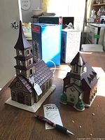 Two bisque ceramic Christmas churches placed on a wooden surface with boxes in the background and a pen to provide scale.