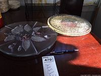 Two clear round glass serving platters placed on a wooden surface, one with purple floral accents and leaf sections, the other with a fish and floral embossed design covered by a glass lid.
