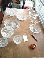 Photo of seven crystal and glass bowls arranged on a wooden table, showing different shapes and cut patterns. Includes measuring tape and auction tag for scale and identification.