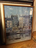 Framed print showing a canal with buildings and boats under glass, leaning against a wall.