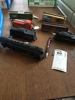 Photo showing HO scale steam locomotive engine, two tenders, three box cars of varied colors and brands, and one orange caboose on a wooden surface with pen for scale.