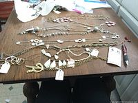 Photo showing a variety of costume necklaces laid out on a wooden table, including gold-tone, silver-tone, white geometric pendants, pink flower accents, and pearl-like beads.