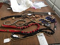 Photo showing various costume necklaces laid out on a wooden surface, including bead styles in black, red, silver, and blue, some with pendants and fabric elements.