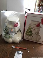 Photo of Lenox snowman cookie jar in styrofoam packaging next to the box depicting the same figure, with a pen for scale.