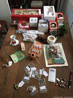 Lot of various Christmas ornaments and decorations displayed on a table including boxed ornaments, jewelry pieces, buttons, candle holder, and wreath hanger.