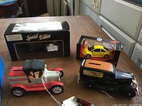 Four boxed diecast model vehicles displayed on wooden table: Jaguar Special Edition in box, yellow VW Beetle in box, plus two antique-style truck banks – red and white 'Ben Franklin' and black 'Gas & Electric' with orange roof.