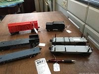 Overview of the five O gauge model train cars arranged on a table: red boxcar, black tender, and three black flatbeds with hooks and ribbed surfaces.
