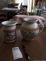 Photo of ceramic spooner and pitcher with floral design on wooden table with pen for scale.