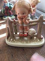 Close-up of ceramic figurine of girl sitting on a rustic wooden fence with sheep detail at base
