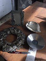 All four items on a wooden table: the round sea motif trivet with shells and seahorses, the metal leaf dish, the small metal coin tray, and the picture frame with ship decorations