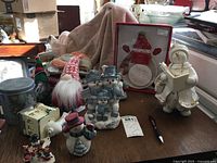 Table showing ceramic and resin snowmen figurines, fabric gnome dolls, Christmas stockings, tin can, and boxed plug-in night light