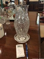 Tall glass candle holder showing diamond cut pattern and flared top, placed on scalloped base plate with a pen for scale.