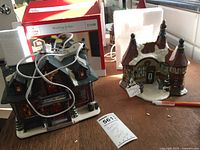 Photo showing both ceramic Christmas village houses side by side, the Hunting Lodge with electrical cord and box, and the Sleigh House with distinct turret design.