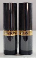 Two Revlon lipstick tubes side by side, black casing with gold band showing Revlon name.