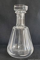 Clear Baccarat Tallyrand cut crystal decanter with matching stopper shown against black background. Vertical cut patterns and geometric stopper facets are visible.