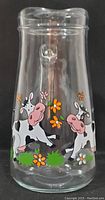 Full view of the clear glass milk pitcher showing the silkscreen decoration of two cartoon cows with flowers and grass.