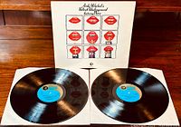 Front view of two vinyl LP records laid flat within the white paper sleeves and the album cover standing behind them, showing the red lips and Coca-Cola bottle graphics.