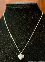 Front view of locket pendant on chain displayed on black velvet stand, showing heart shape and scrollwork decoration.