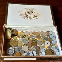 Open wooden Sueno Panza Habana box containing a collection of mixed coins of various sizes and metals spread inside the box.