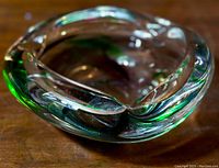 Angled top view of the thick clear glass ashtray with green colored band inside. Smooth curved edges and three cigarette rests visible.