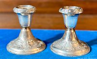 Pair of ornate sterling silver weighted candlesticks shown on blue cloth, highlighting detailed scrollwork around base and rim.