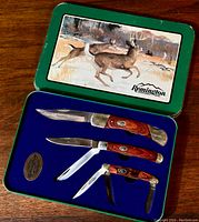 Set of four folding knives displayed in open green tin case with lid showing deer artwork on outside and blue velvet interior lining.