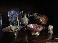 Shelf display with the mixed lot items grouped, including brass bowls, etched brass vase, glass vase with marbles, elephant figurine, and incense burner
