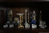 Shelf display of multiple assorted candle holders including brass and glass pieces along with two oil lamps