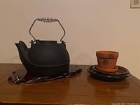 Full view of cast iron kettle on wooden trivet next to terracotta planter and drip trays
