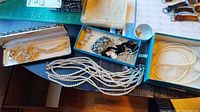 Photo showing multiple faux pearl necklaces on a table along with a box of assorted jewelry including earrings and brooches.