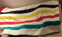 Multicolor striped wool Hudson Bay blanket folded on couch showing stripes in black, yellow, red, and green on cream background.