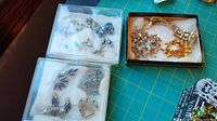 Jewelry pieces displayed in small transparent and black boxes on a green cutting mat, showing various earrings, bracelets, and brooches.