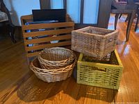 Wooden magazine rack holding several magazines and five assorted woven baskets grouped beside it on a hardwood floor.