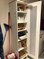 Cabinet with open door revealing shelves filled with envelopes, folders, and paper stacks. Cleaning tools visible beside the cabinet.