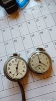 Photo showing two Westclox pocket watches on a calendar sheet, displaying faces and winding crowns.