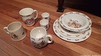 Set of Royal Doulton Bunnykins dishes including one large mug, two small cups, creamer jug, two bowls, and four plates arranged on a wooden floor.