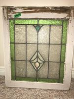 Whole stained glass window panel upright against a wall, showing full design of textured clear glass with green border and central diamond motif with amber and turquoise glass.