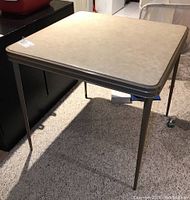 Front side angle view of the square folding table with vinyl top and metal legs standing on carpet.