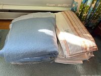 Two blankets, one blue and one white with beige stripes, stacked side by side on a greenish carpet near a radiator and floral curtains.