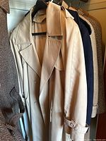 Photo showing several women's coats and jackets hanging, focusing on light beige trench coat with double-breasted button front.