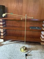 Full view of the adjustable height floor lamp leaning on a wooden shelf, showing the brass finish, base, arm, and black foot pedal.