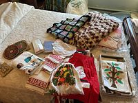 Photo showing assortment of kitchen linens including crocheted afghans, aprons, embroidered towels, trivets, and linens laid out on a bed.